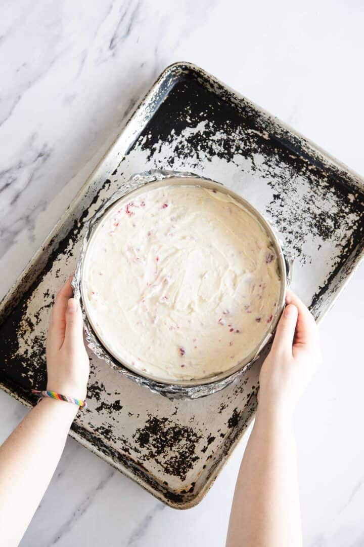 A person places a round cake pan filled with light-colored, berry-speckled Raspberry Ricotta Cheesecake batter onto a worn, dark baking sheet on a marble surface.