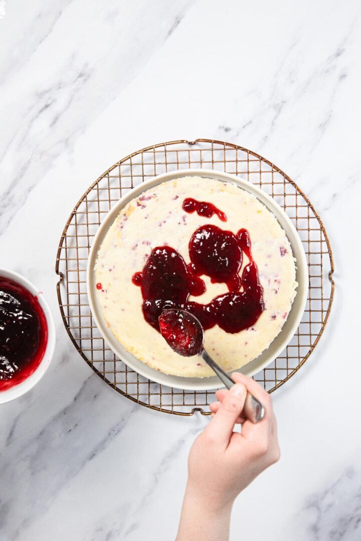 A hand spreads a spoonful of red fruit sauce over a Raspberry Ricotta Cheesecake on a wire rack, with a bowl of extra sauce nearby on the white marble surface.