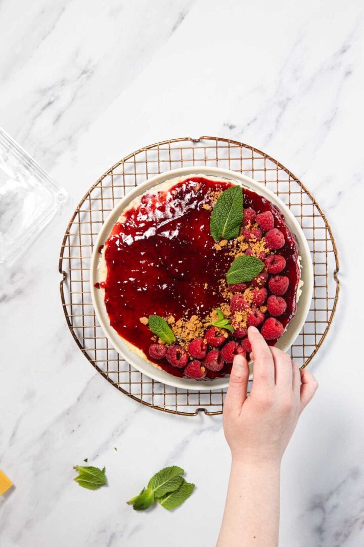 A hand places a mint leaf on a Raspberry Ricotta Cheesecake topped with raspberry sauce, fresh raspberries, mint leaves, and crumb topping, set on a wire rack over a marble surface.