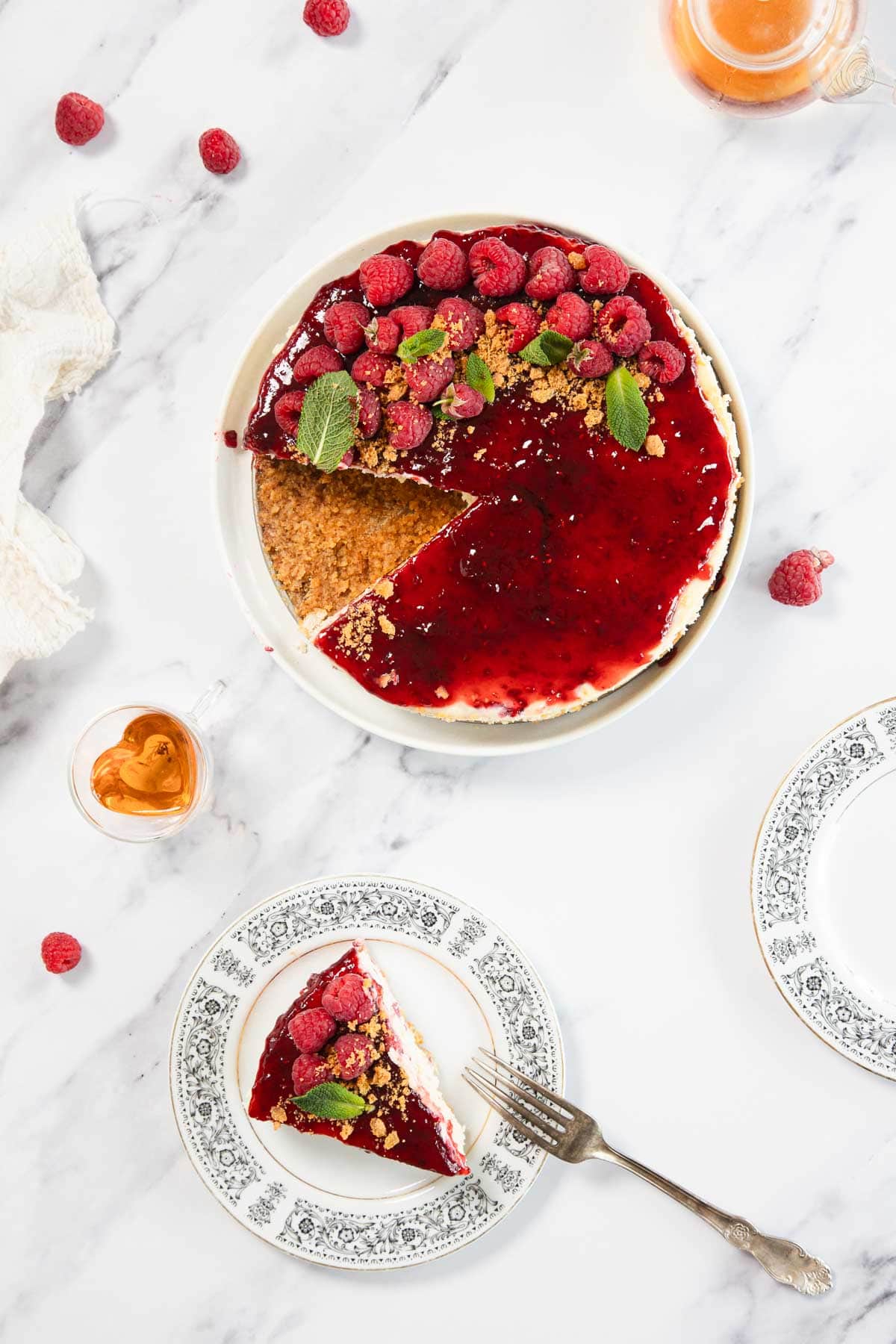 A Raspberry Ricotta Cheesecake with red raspberry topping, garnished with fresh raspberries and mint leaves, sits on a marble surface. One slice is on a patterned plate with a fork; empty plate, raspberries, and drink nearby.