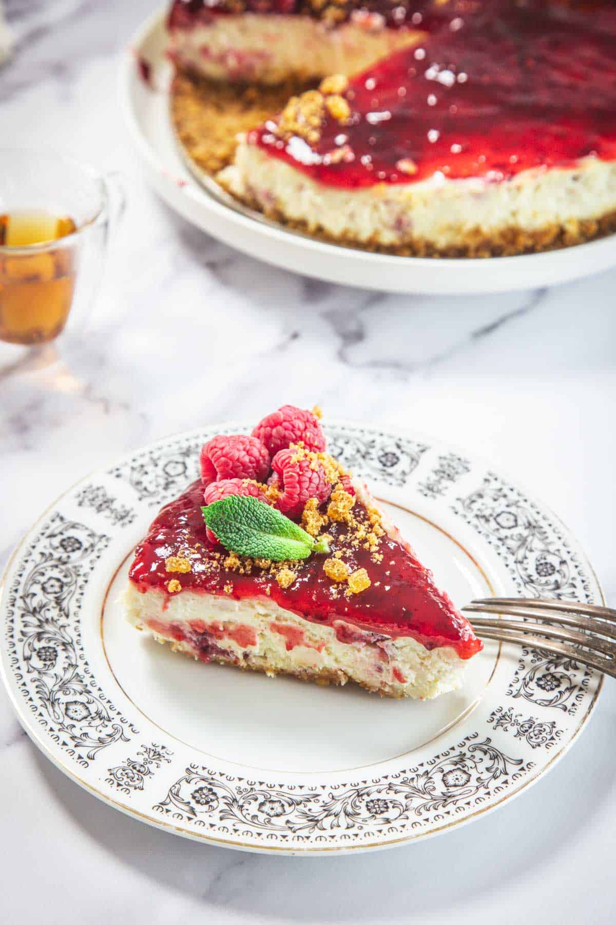 A slice of Raspberry Ricotta Cheesecake with raspberry topping, fresh raspberries, and mint leaves sits on a patterned plate, fork beside it. The rest of the cheesecake is visible in the background on a white platter.