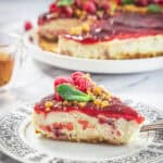 A slice of Raspberry Ricotta Cheesecake with a red glaze, garnished with fresh raspberries and mint, sits on a decorative plate; the rest of the cheesecake is visible in the background.