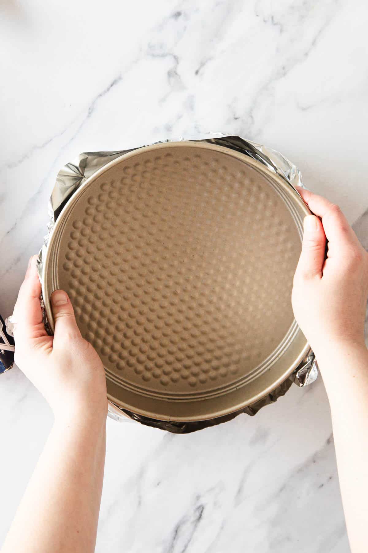 Two hands hold a round, textured-bottom cake pan wrapped in aluminum foil on a white marble surface—perfect for baking a Raspberry Ricotta Cheesecake.
