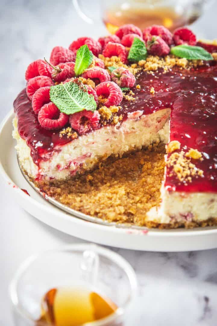 A Raspberry Ricotta Cheesecake with a golden brown crumb crust, topped with raspberry sauce, fresh raspberries, and mint leaves. A slice is removed to reveal the creamy interior. A glass of tea sits nearby.