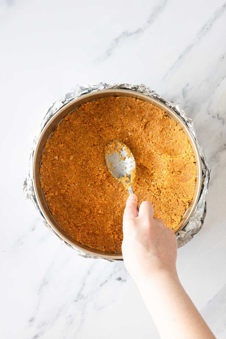 A hand uses a spoon to press down a crumbly crust mixture for Raspberry Ricotta Cheesecake in a foil-wrapped round springform pan on a white marble surface.