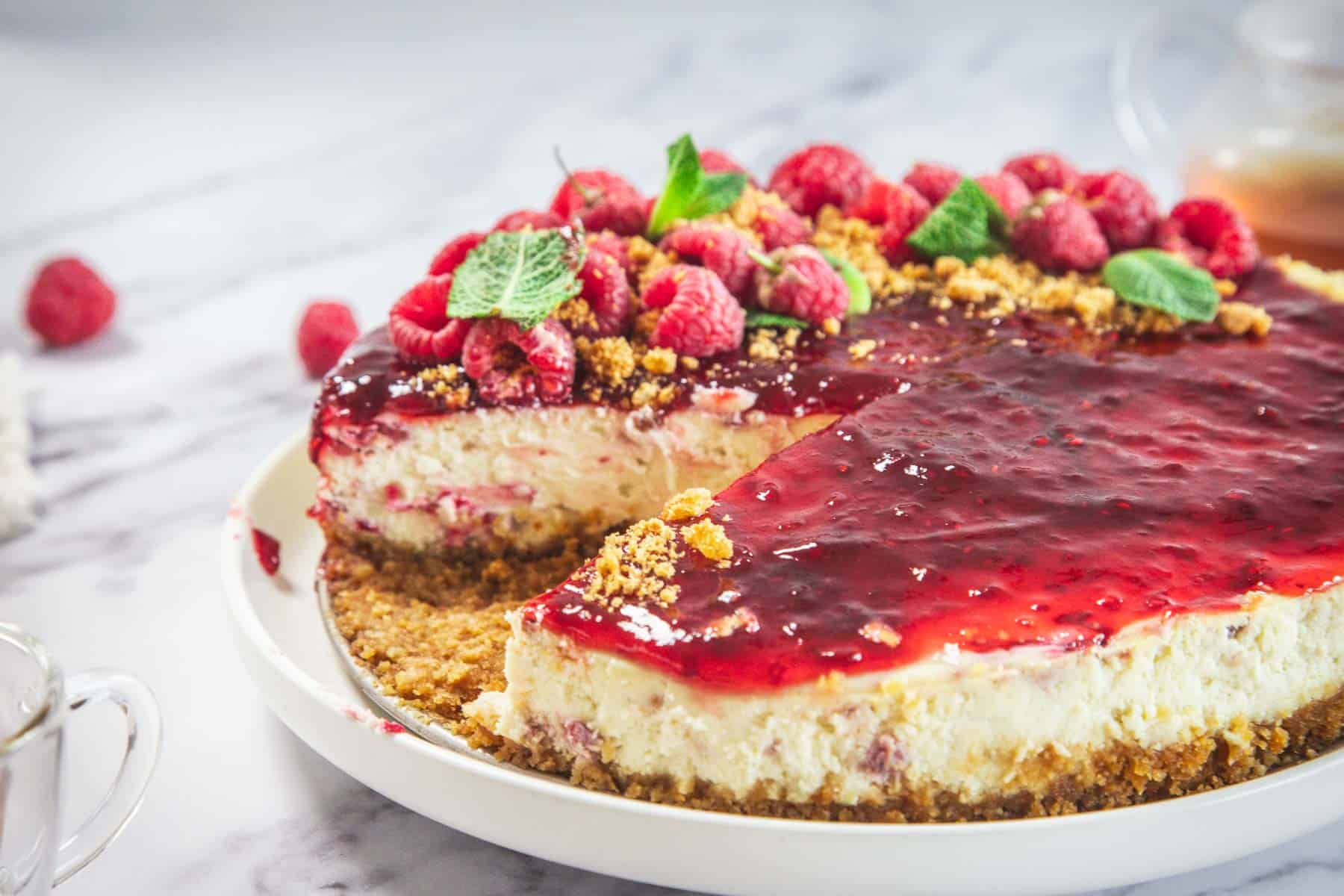 A Raspberry Ricotta Cheesecake with a graham cracker crust, topped with raspberry sauce, fresh raspberries, mint leaves, and crumbled topping. A large slice is missing, revealing the ultra-creamy ricotta interior.