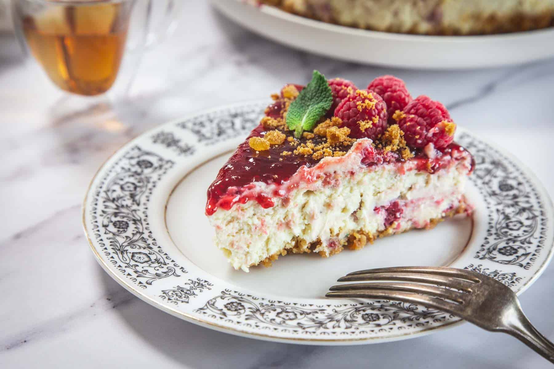 A slice of Raspberry Ricotta Cheesecake topped with raspberry sauce, fresh raspberries, crumbled cookie, and a mint leaf sits on a decorative plate with a fork. A glass of tea and more cheesecake are in the background.