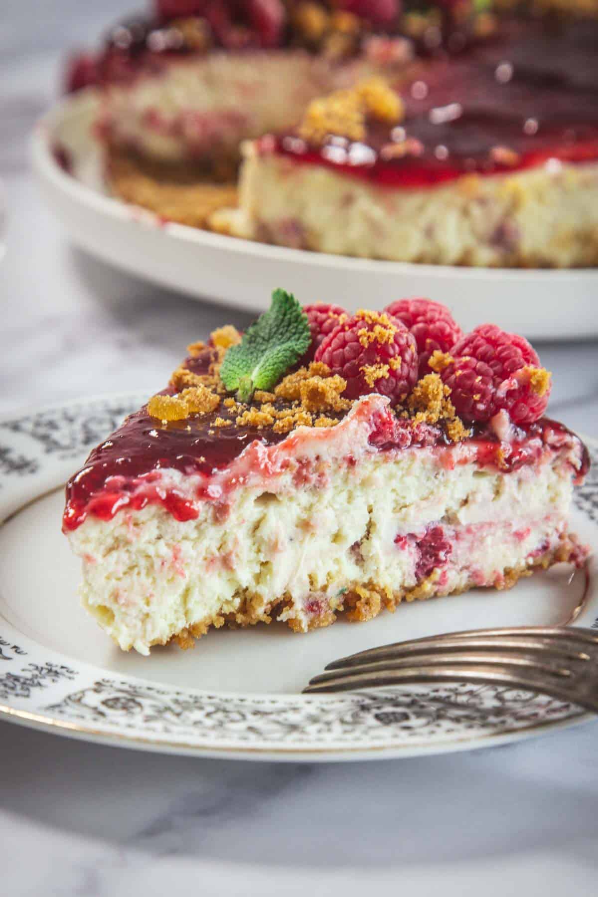 A slice of Raspberry Ricotta Cheesecake with a crumbly crust, raspberry topping, fresh raspberries, and a mint leaf sits on a patterned plate with a fork. The rest of the cheesecake is visible in the background.