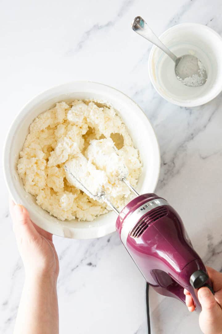 A person holds a white bowl of creamed mixture—perfect for Raspberry Ricotta Cheesecake—while using a purple hand mixer. Nearby, a small bowl with a spoon and white powder sits on the marble countertop.