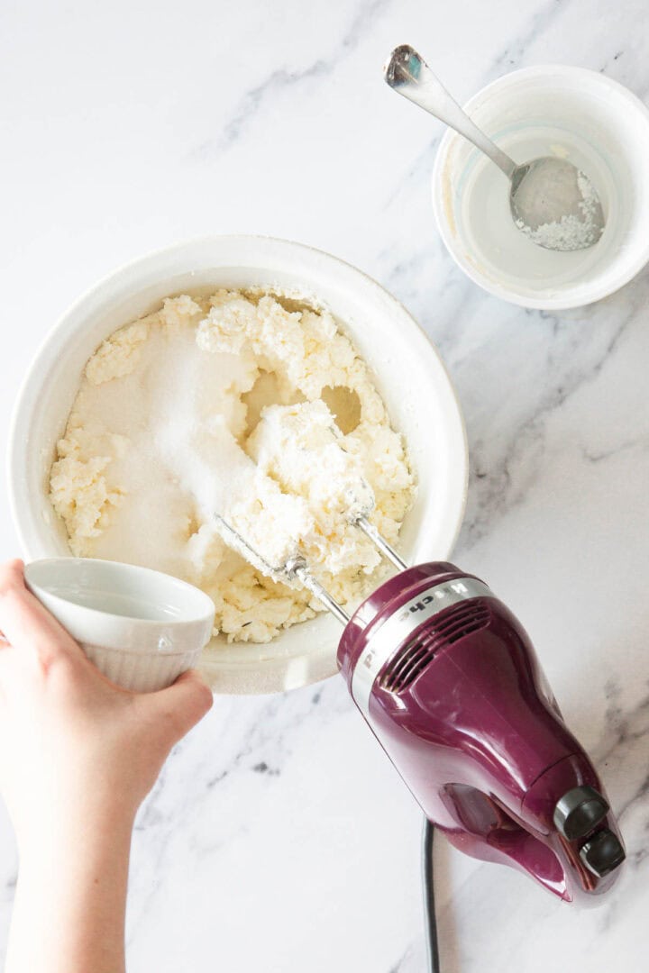 A person pours liquid from a small white cup into a mixing bowl with sugar and butter for a Raspberry Ricotta Cheesecake, while using a hand mixer. Two white bowls and a spoon rest on the marble countertop.