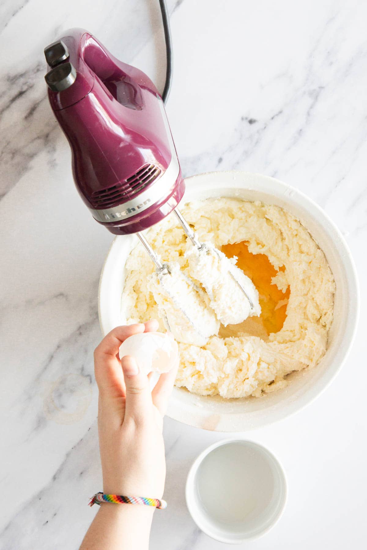 A hand cracks an egg into a white mixing bowl filled with creamed ingredients for Raspberry Ricotta Cheesecake. A purple electric hand mixer rests over the bowl, and a small empty bowl sits nearby on a marble surface.