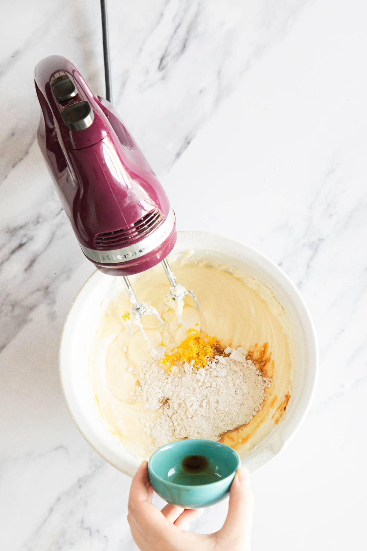A hand holds a small turquoise bowl above a mixing bowl filled with batter, flour, and orange zest for Raspberry Ricotta Cheesecake, while a maroon electric hand mixer rests nearby on the white marble surface.