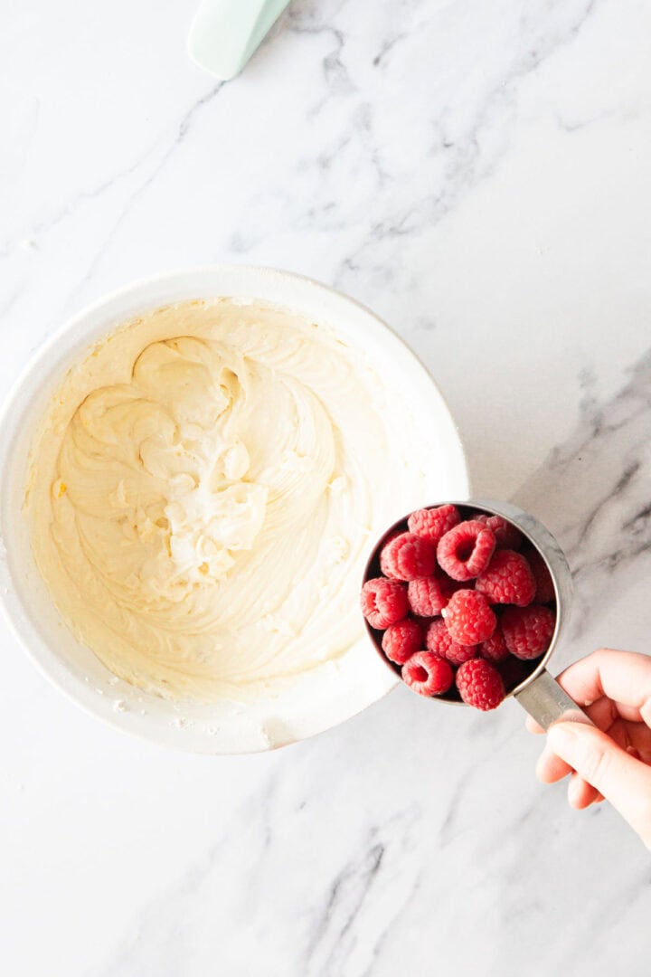 A hand holds a metal measuring cup filled with fresh raspberries over a white bowl of creamy batter—perfect for making Raspberry Ricotta Cheesecake—on a marble countertop.