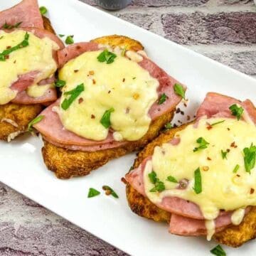 Two slices of toasted bread topped with ham, melted cheese, and chopped parsley are on a white rectangular plate. They rest on a brick-patterned surface, with part of a floral-patterned cloth and a bowl of diced tomatoes visible in the background.