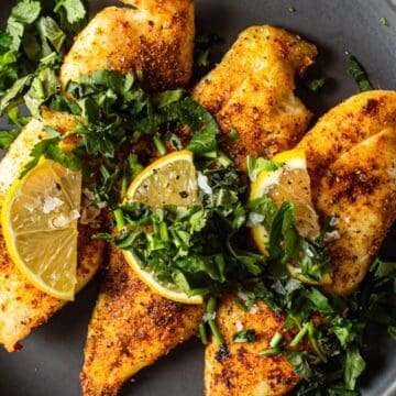 Four pieces of seasoned, cooked chicken breast on a gray plate, garnished with chopped fresh herbs and lemon wedges, placed on a white marble surface.