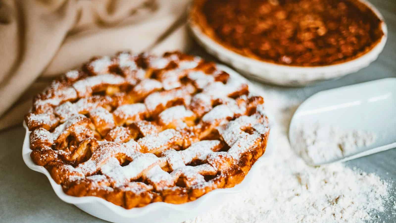 Two pies on a table: a front pie with a lattice top dusted with powdered sugar and a second pie in the background, accompanied by a scoop with some spilled flour. A light cloth lies partly visible on the table.