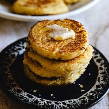 A stack of three golden-brown Arepas de Choclo sits on a black speckled plate, topped with a pat of melting butter. A few crumbs are scattered around, and more patties are visible on a plate in the background.