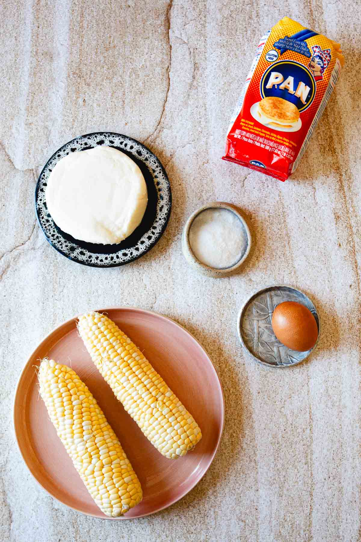 A round plate with two ears of corn, a black and white plate with a round block of cheese, a whole egg, a small dish of salt, and a package of PAN corn flour are arranged on a light-colored surface—perfect for making Arepas de Choclo.