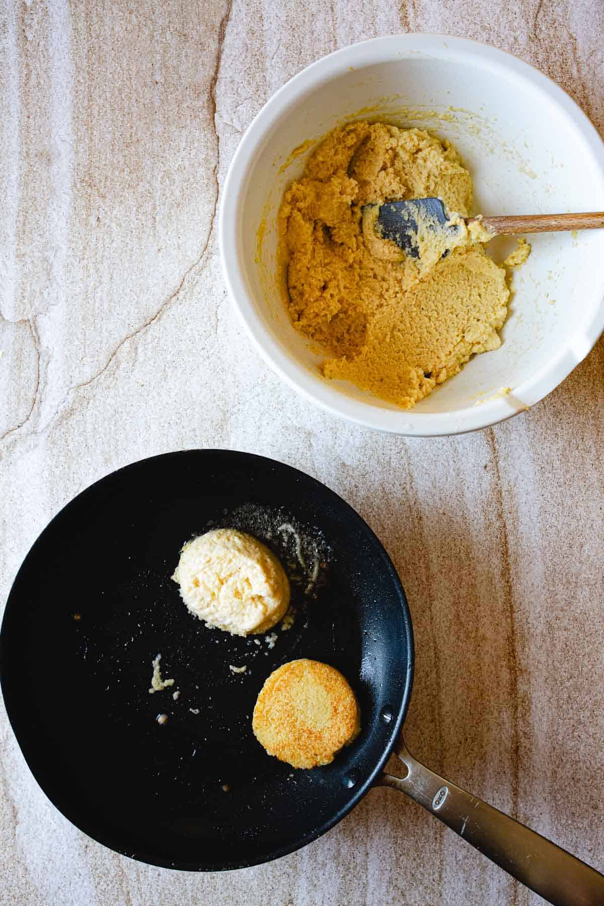 A black frying pan with two Arepas de Choclo, one uncooked and one partially cooked, sits on a countertop next to a white bowl containing yellow batter and a spatula.