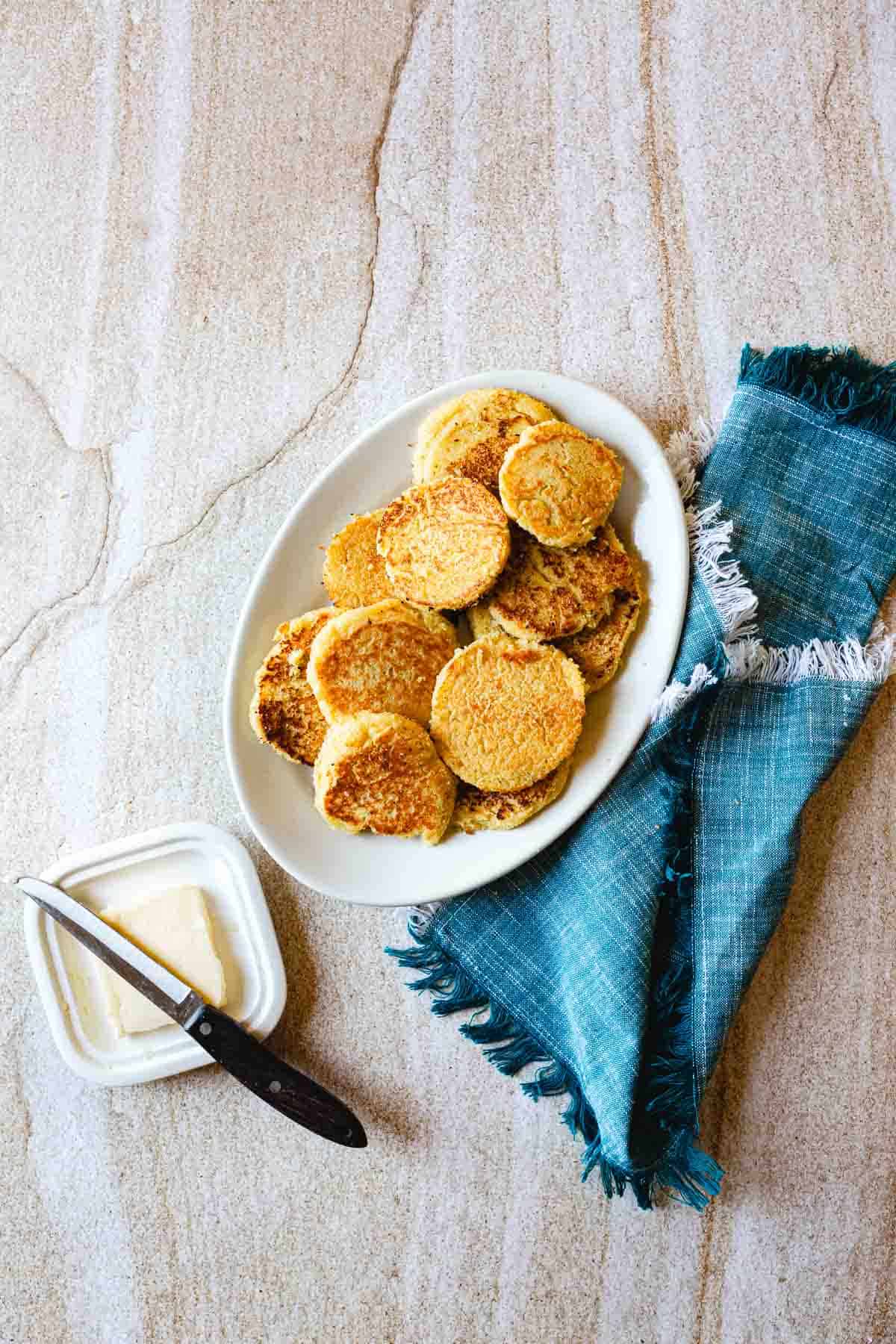 A white oval plate with a stack of small round Arepas de Choclo sits on a stone surface next to a blue fringed napkin. Nearby, a container of butter with a knife resting on top completes the inviting scene.