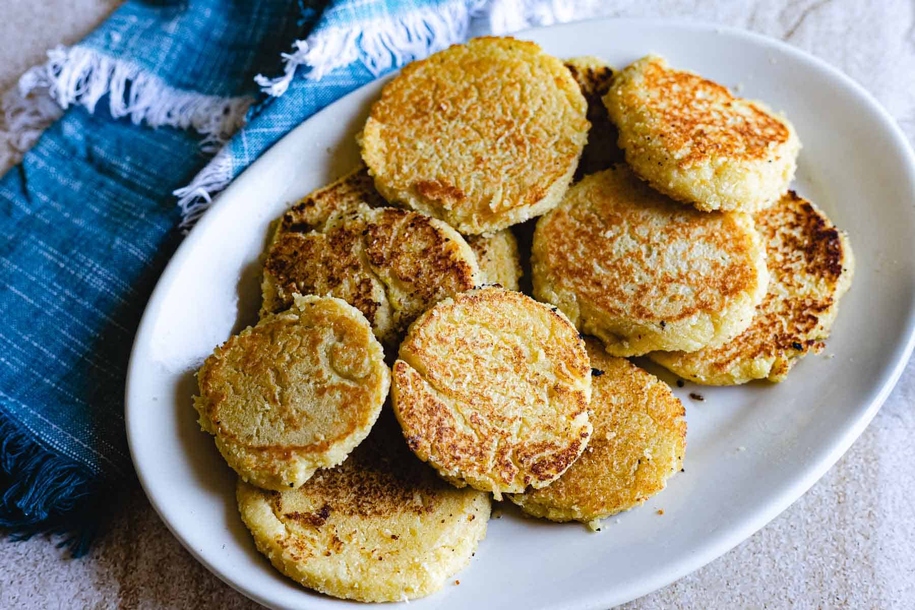 A white oval plate holds several golden-brown, round Arepas de Choclo. The plate rests next to a blue cloth napkin with frayed edges on a light-colored surface.