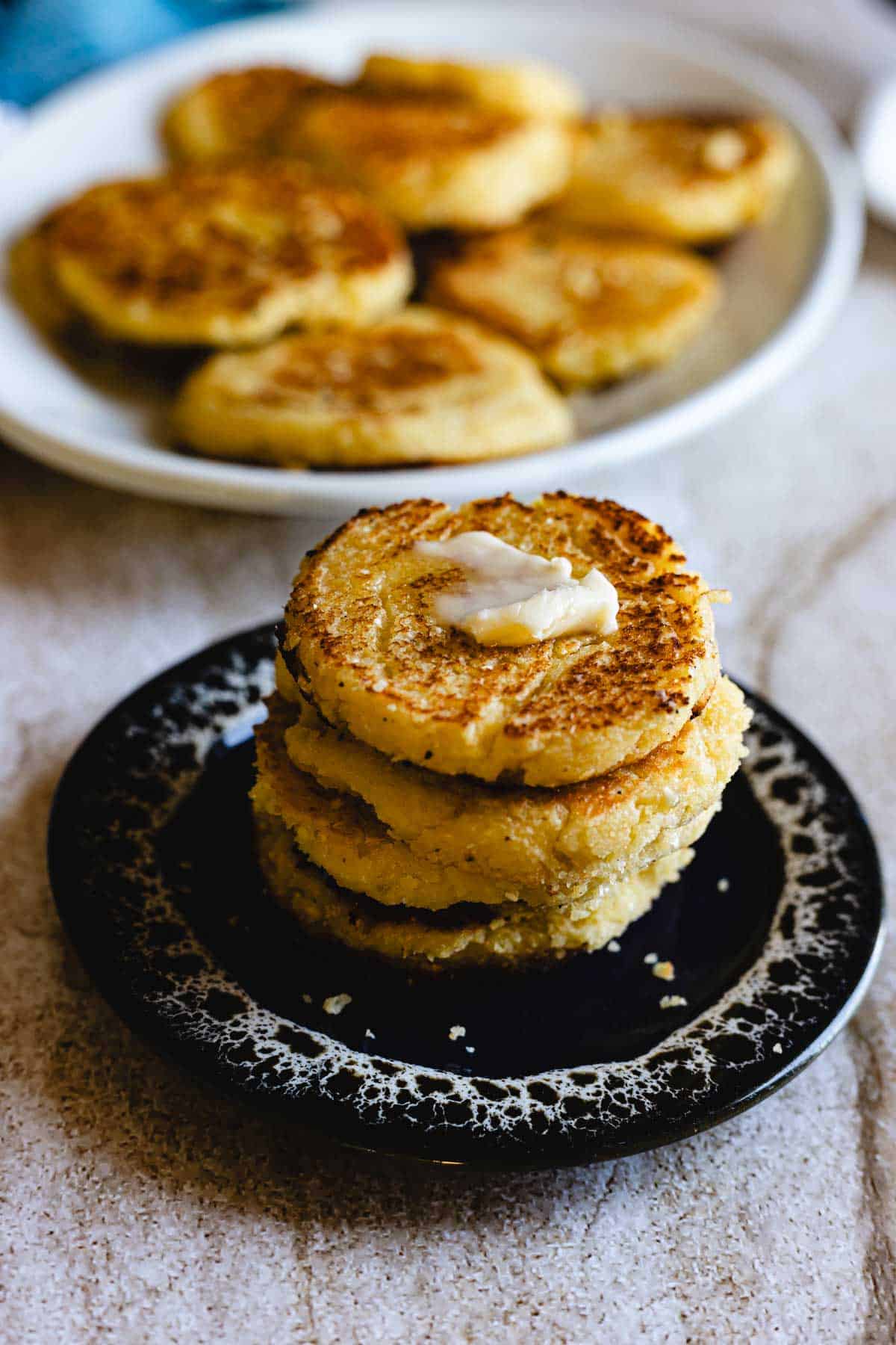 Three golden brown Arepas de Choclo, round cornmeal cakes, are stacked on a small dark plate with a pat of butter melting on top, while more cornmeal cakes await in the background on a white plate.
