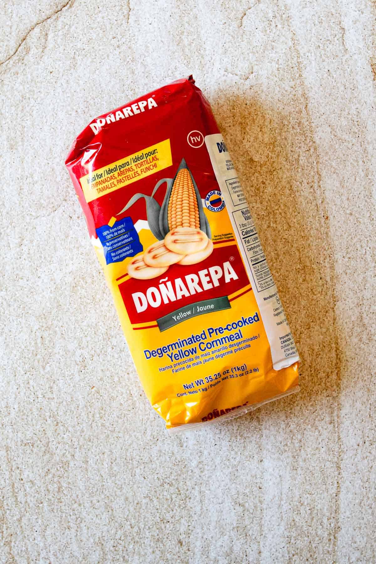 A package of Doñarepa Degerminated Pre-cooked Yellow Cornmeal lies on a beige surface. The red and yellow packaging features an image of corn on the cob and round corn patties, perfect for making traditional Arepas de Choclo.