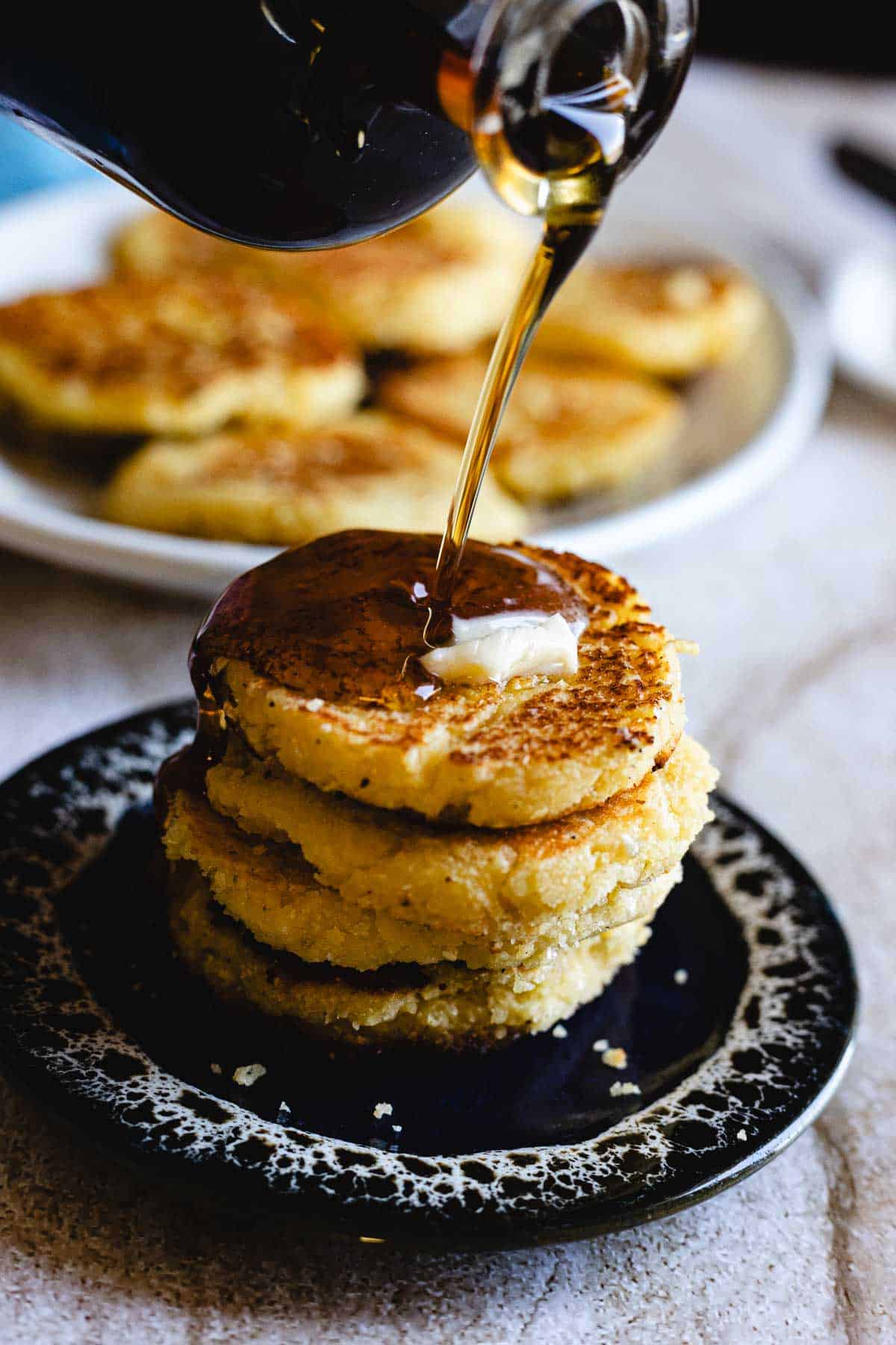 A stack of four Arepas de Choclo, golden and fluffy, sits on a black plate topped with butter as syrup is poured over them. In the background, more Arepas de Choclo rest temptingly on a white plate.