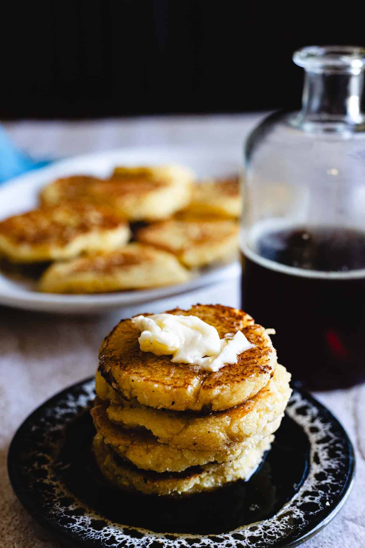 A stack of Arepas de Choclo, also known as cornbread cakes, topped with a pat of butter sits on a dark plate. In the background, more Arepas de Choclo and a glass bottle of syrup are visible.