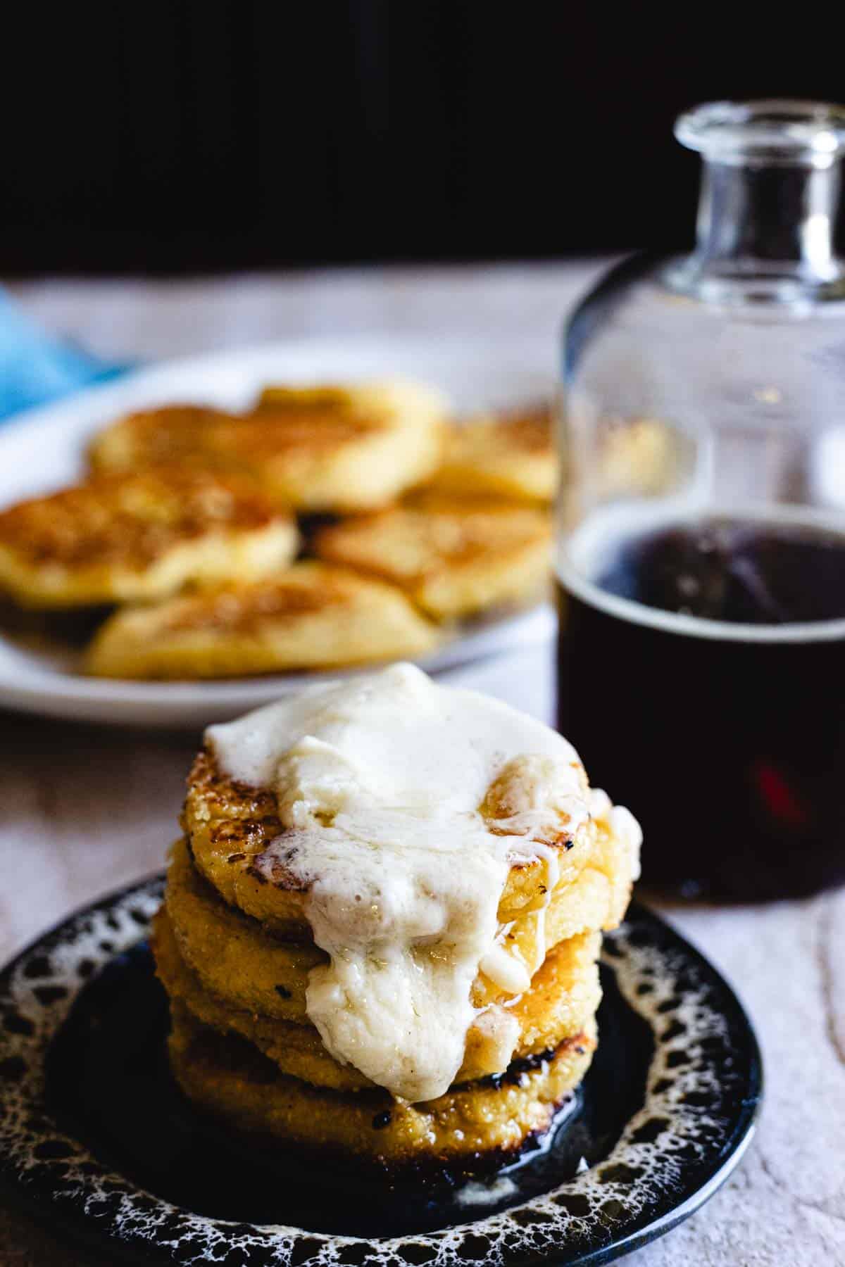 A stack of Arepas de Choclo, golden fried cornbread cakes topped with melted cheese, sits on a dark plate. In the background, a glass bottle with a dark liquid and a plate with more arepas are visible.