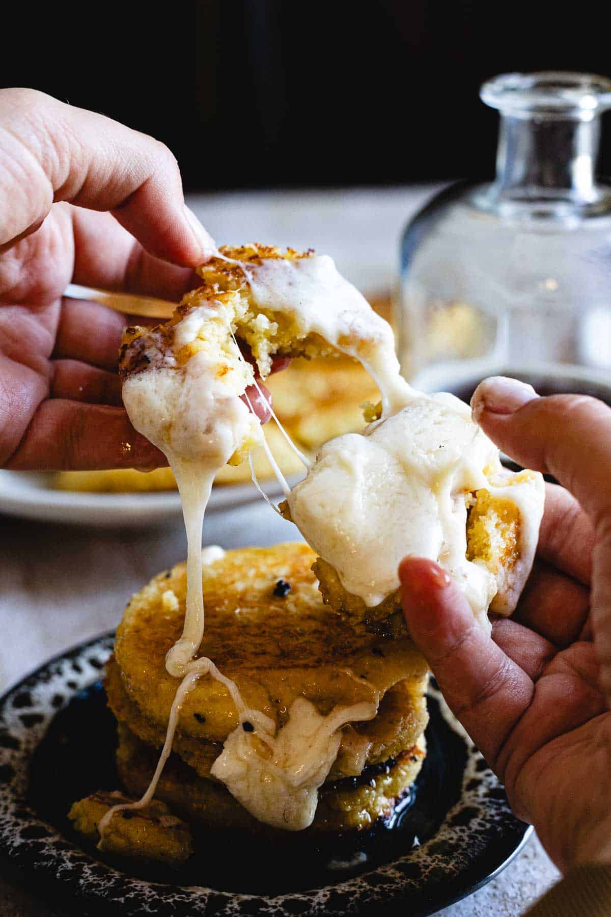 A person pulls apart a stack of Arepas de Choclo filled with melted, stretchy cheese, revealing the gooey center. The golden patties are served on a patterned plate, with a glass jar in the background.