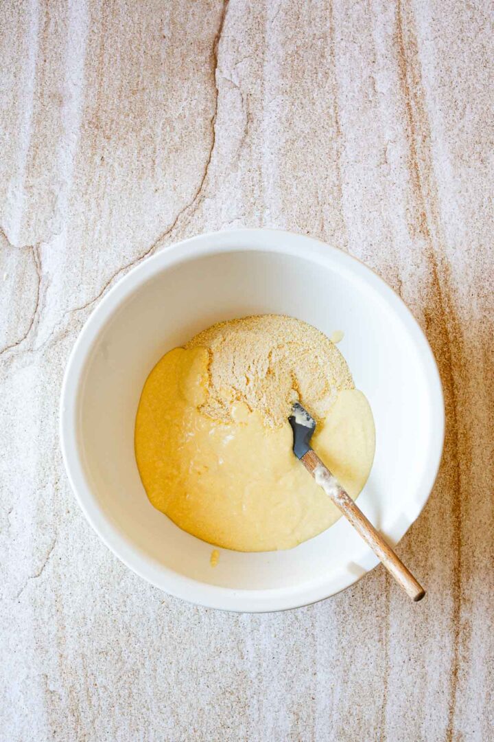 A white mixing bowl containing a thick yellow Arepas de Choclo batter and a pile of dry ingredients sits on a light-colored countertop. A spatula with a wooden handle is resting in the bowl.