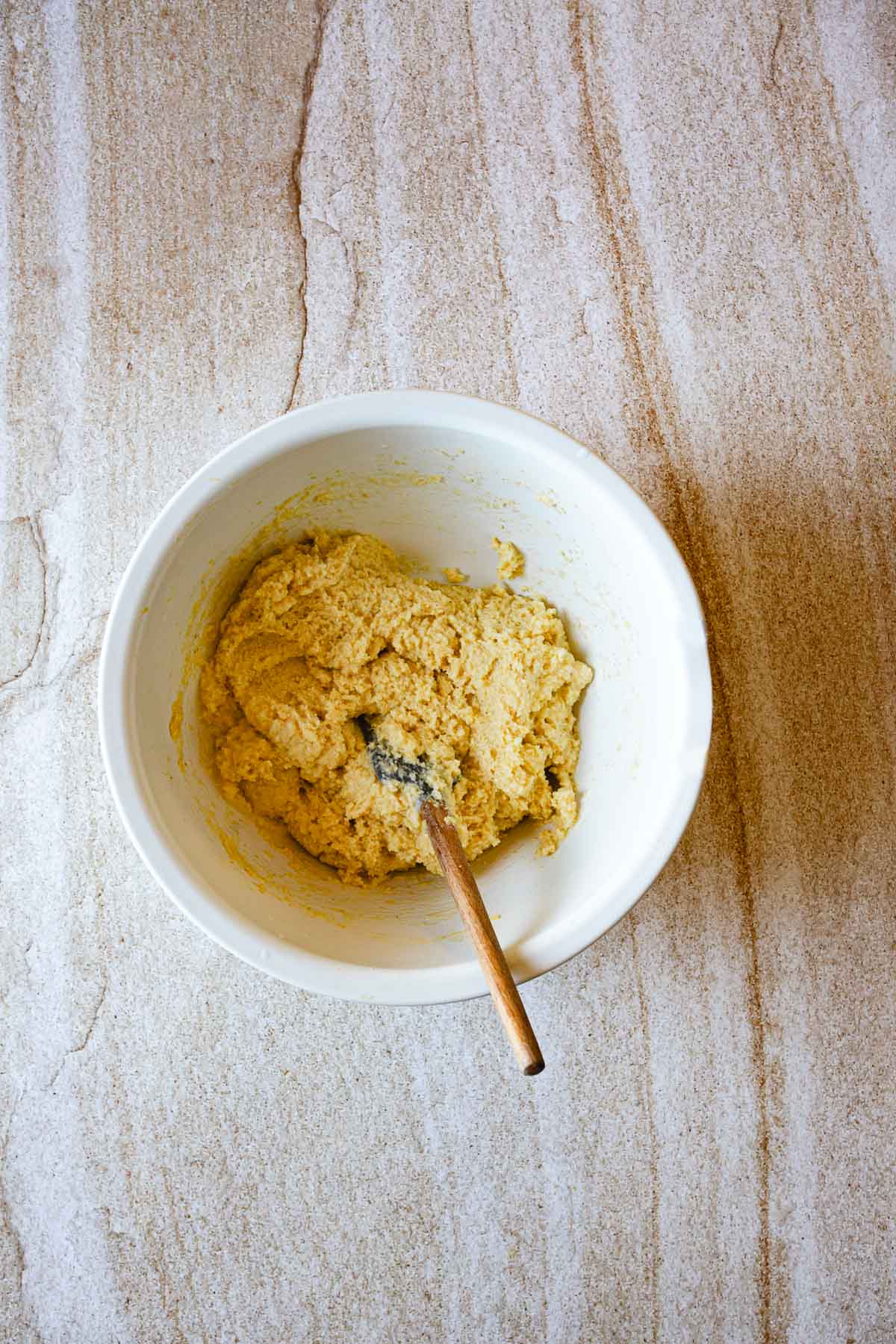 A white mixing bowl containing a thick, yellowish Arepas de Choclo dough sits on a light-colored, marbled countertop. A wooden spoon with a black tip is resting inside the bowl.
