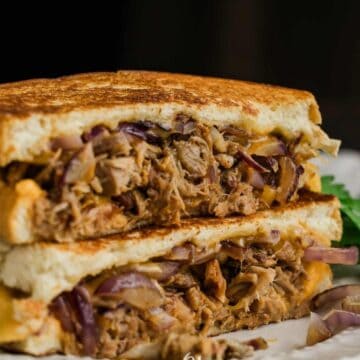 A close-up of an all-time favorite sandwich, cut in half and stacked. It contains shredded meat, red onions, and cheese, with grill marks on the bread. A piece of cilantro sits next to this delicious creation on a white plate, a nod to timeless favorite recipes enjoyed by boomers everywhere.