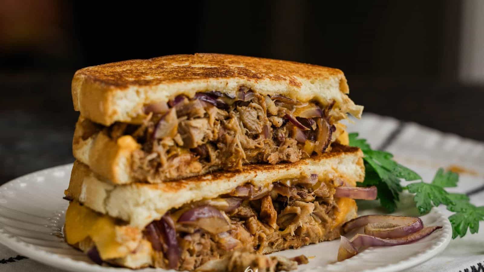 A close-up of an all-time favorite sandwich, cut in half and stacked. It contains shredded meat, red onions, and cheese, with grill marks on the bread. A piece of cilantro sits next to this delicious creation on a white plate, a nod to timeless favorite recipes enjoyed by boomers everywhere.