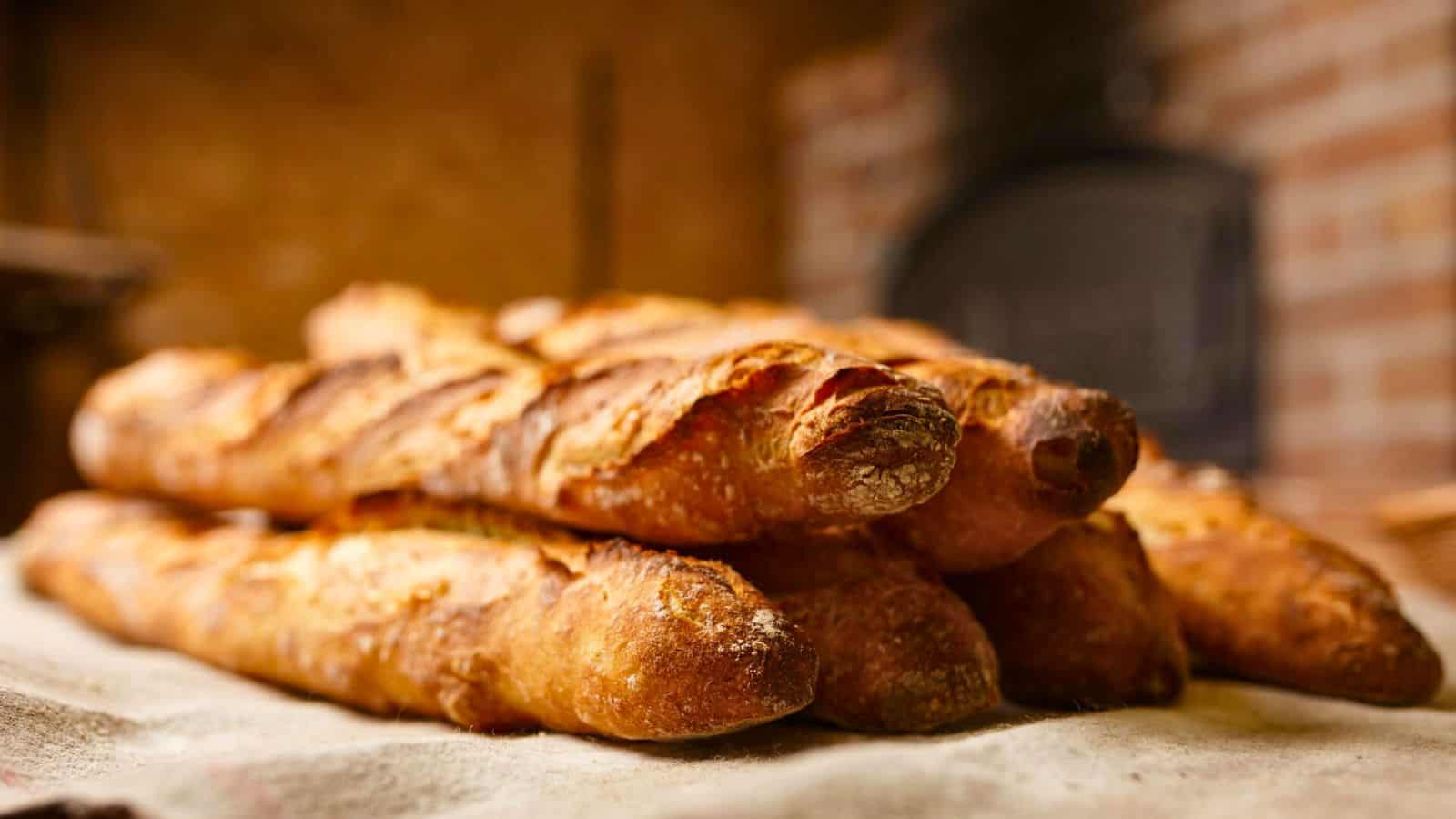 A stack of four freshly baked baguettes rests on a cloth surface, their golden-brown crust crackling with promise. A brick oven blurs in the background, evoking a rustic bakery setting—reminding us that baguettes are among the 13 foods people think are American but aren't.