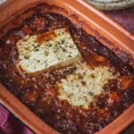 A rectangular baking dish contains baked feta on a bed of cherry tomatoes and Mexican salsa, seasoned with black pepper and herbs. The edges of the dish show some caramelization, adding depth to this savory blend of baked feta in salsa.