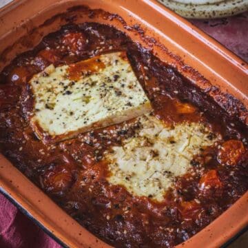 A rectangular baking dish contains baked feta on a bed of cherry tomatoes and Mexican salsa, seasoned with black pepper and herbs. The edges of the dish show some caramelization, adding depth to this savory blend of baked feta in salsa.