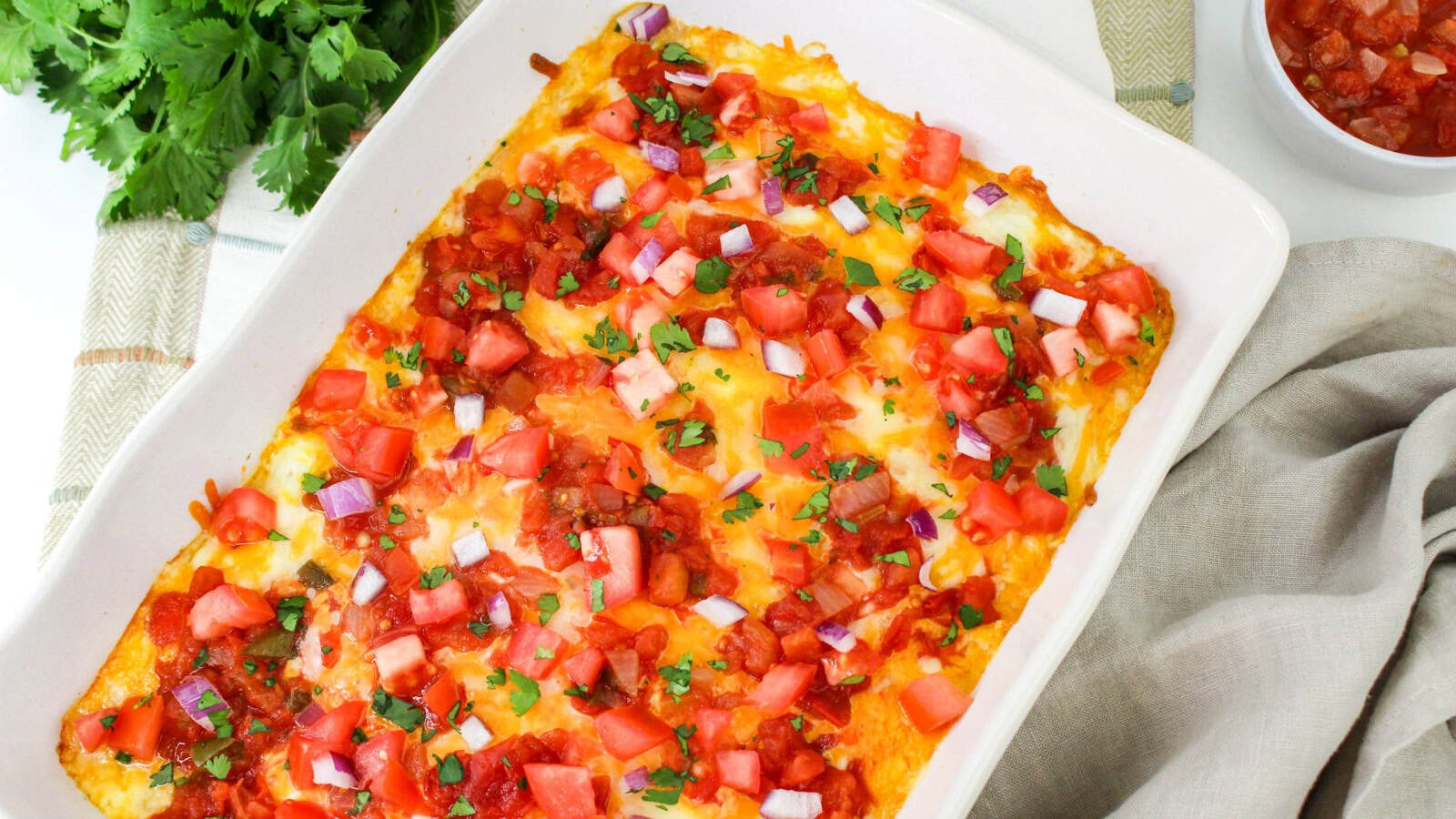 Casserole dish with melted cheese, diced tomatoes, onions, and chopped cilantro on top, with a side bowl of salsa and cilantro garnish nearby.
