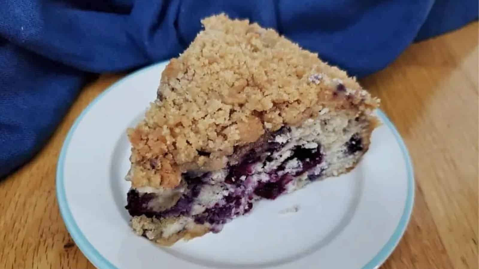 A slice of crumb-topped berry coffee cake sits on a white plate with a blue rim, perfect for Mother’s Day desserts, positioned on a wooden table with a dark blue cloth in the background.