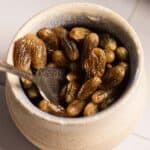 A ceramic jar filled with brandy-soaked raisins cradles a metal spoon that rests against its inside. The jar is placed on a tiled surface, promising a rich blend of flavors within.