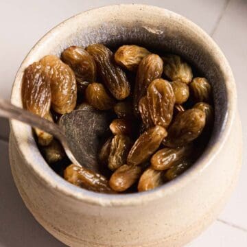 A ceramic jar filled with brandy-soaked raisins cradles a metal spoon that rests against its inside. The jar is placed on a tiled surface, promising a rich blend of flavors within.