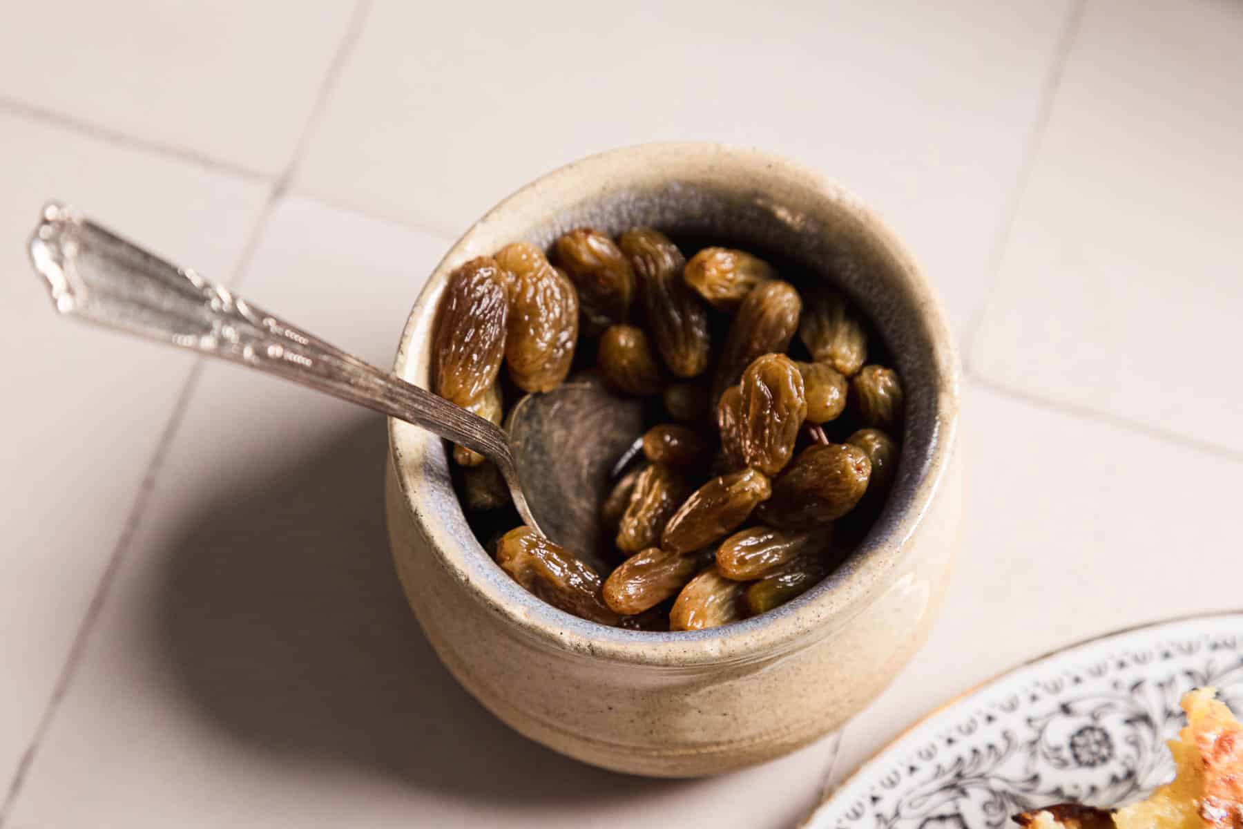 A ceramic bowl brimming with brandy-soaked raisins and a silver spoon rests inside. The bowl is placed on a tiled surface, with part of a decorative plate peeking next to it.