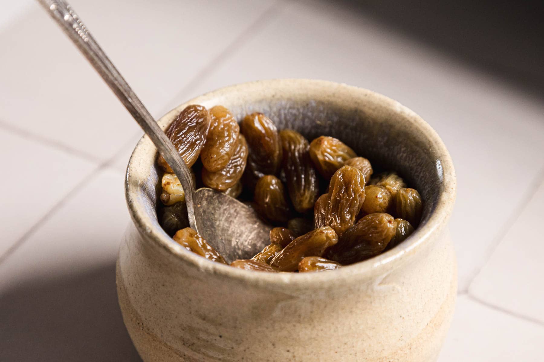 A small ceramic bowl filled with luscious soaked raisins sits on a tiled surface. A silver spoon rests inside, partially submerged in the plump fruit. The natural lighting casts gentle shadows across the bowl, evoking a warm and inviting scene.