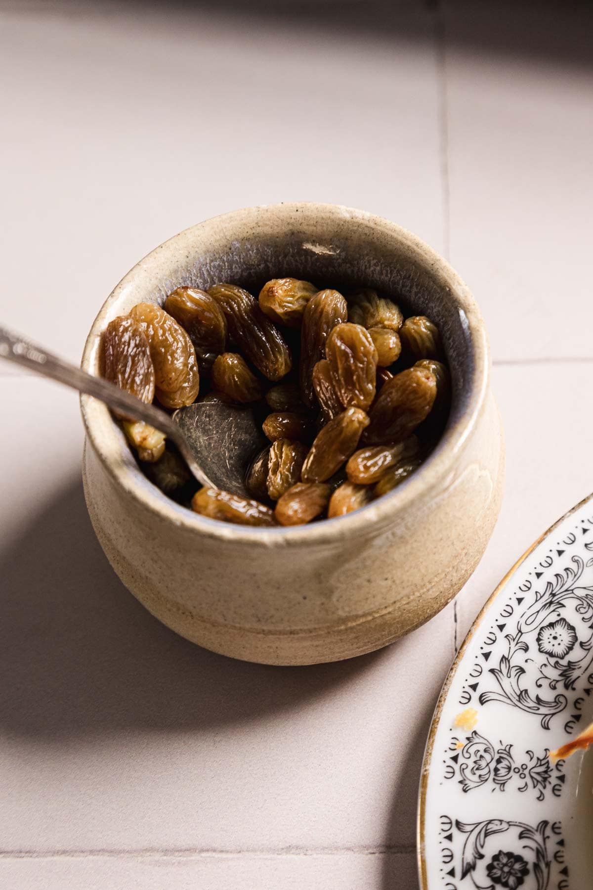 A ceramic bowl filled with golden raisins sits on a beige surface. A metal spoon is partially submerged in the raisins. Part of a decorative black and white plate is visible in the bottom right corner.