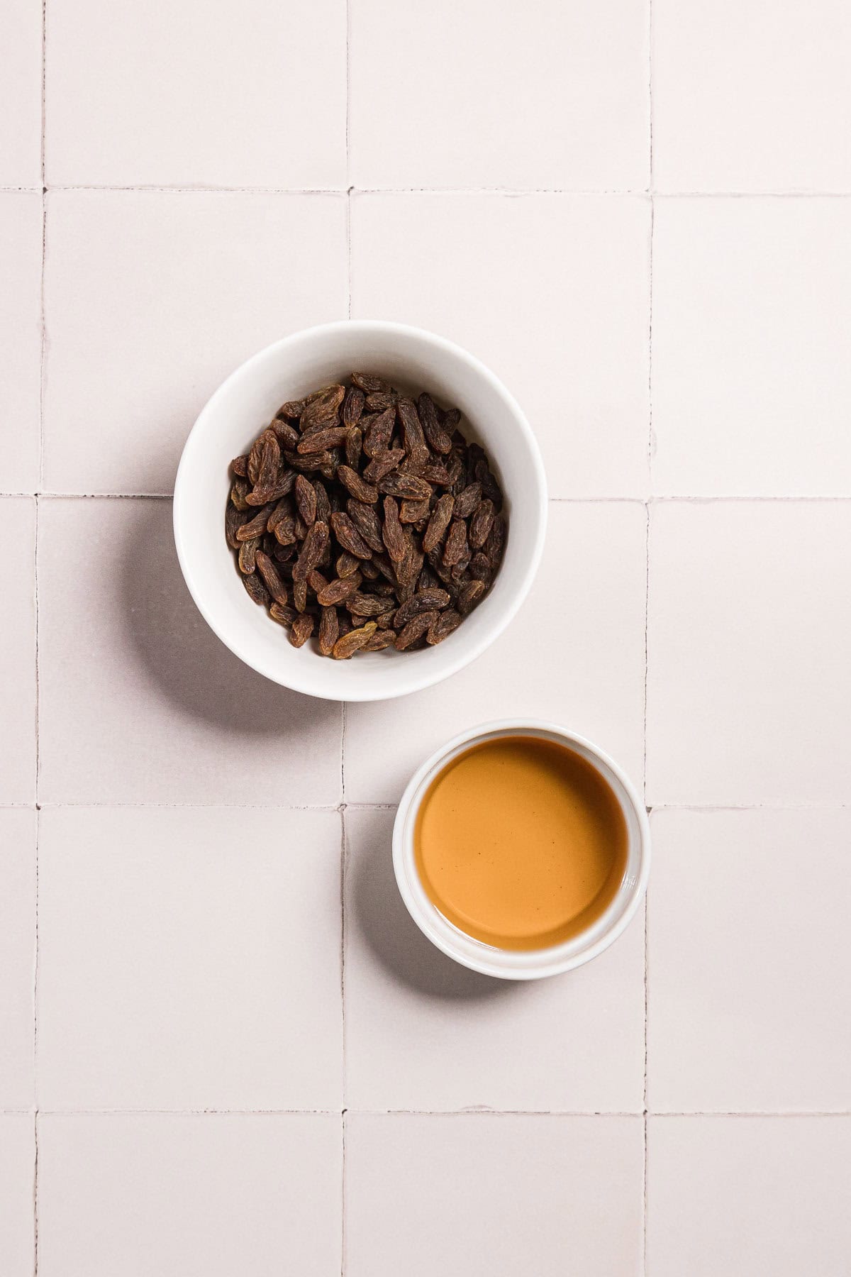 A small white bowl filled with raisins sits next to a small white cup containing a light brown liquid, both placed on a tiled surface.