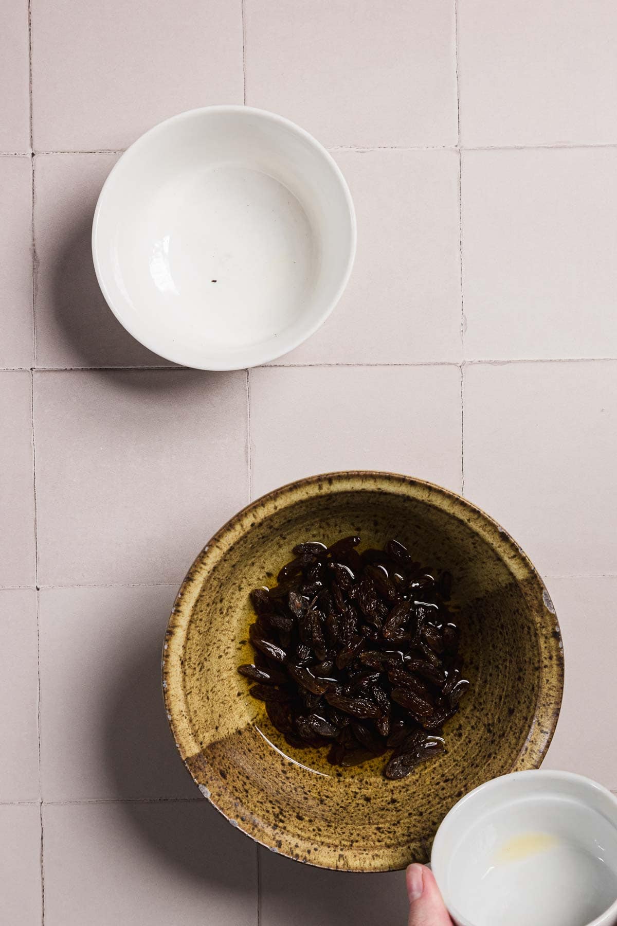 A hand holds a small white bowl above a larger brown bowl filled with prunes in syrup. Another empty white bowl is in the top left corner. The surface beneath is a light tiled countertop.
