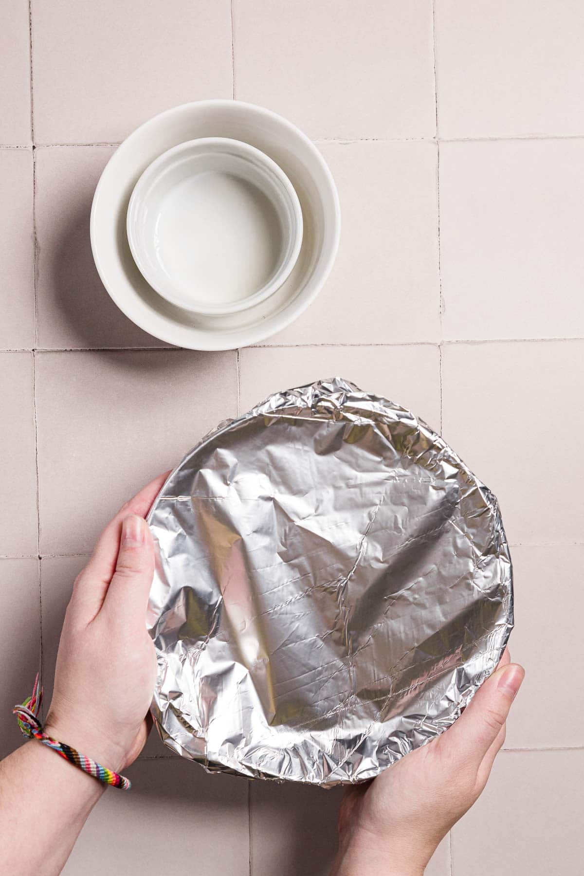 Hands holding a round dish covered in aluminum foil, with two nested white ceramic bowls placed on a tiled surface above. A woven bracelet can be seen on one wrist.