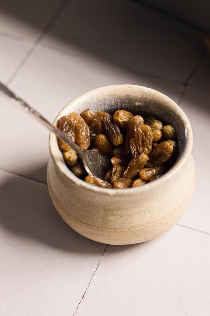 A ceramic bowl filled with dried raisins sits on a tiled surface. A metal spoon is resting inside the bowl. The scene is lit by natural light from the side.