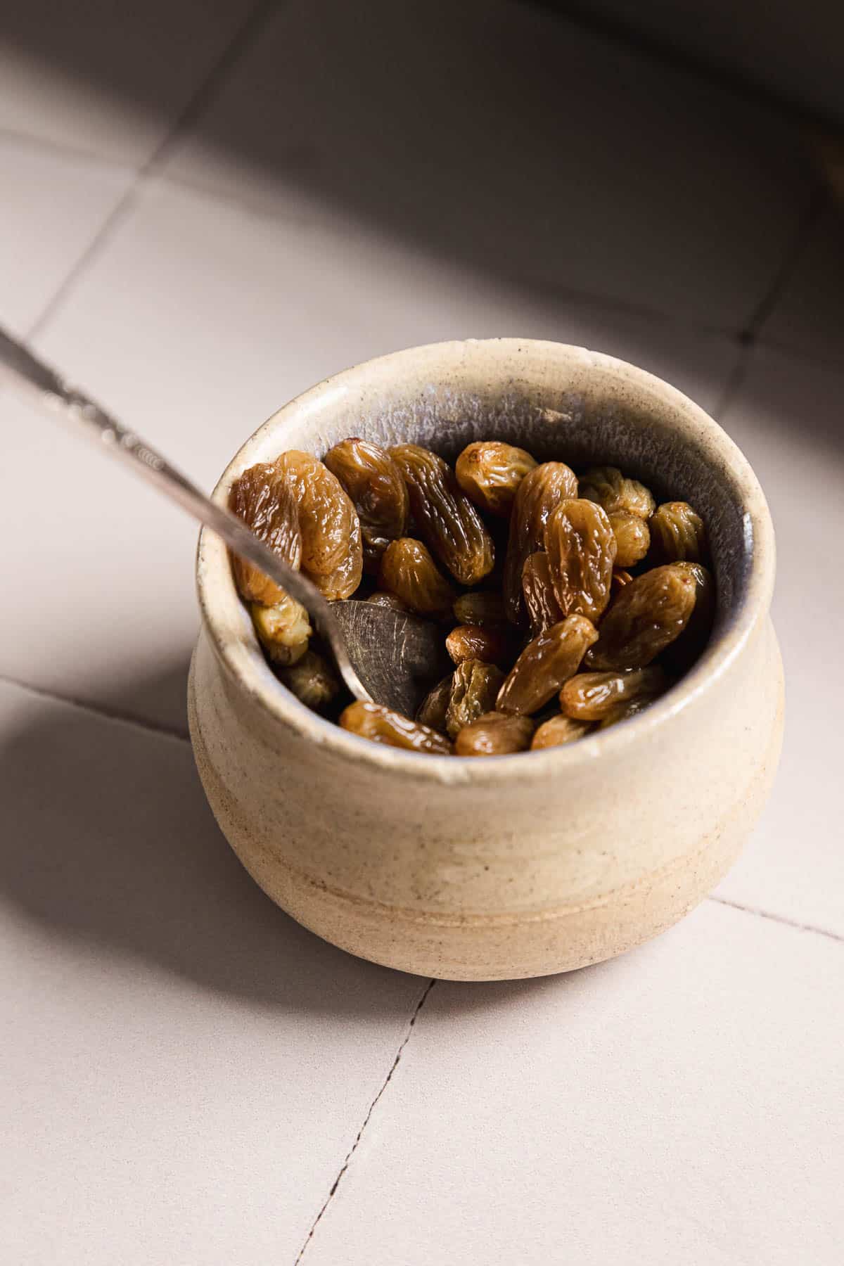 A ceramic bowl filled with dried raisins sits on a tiled surface. A metal spoon is resting inside the bowl. The scene is lit by natural light from the side.